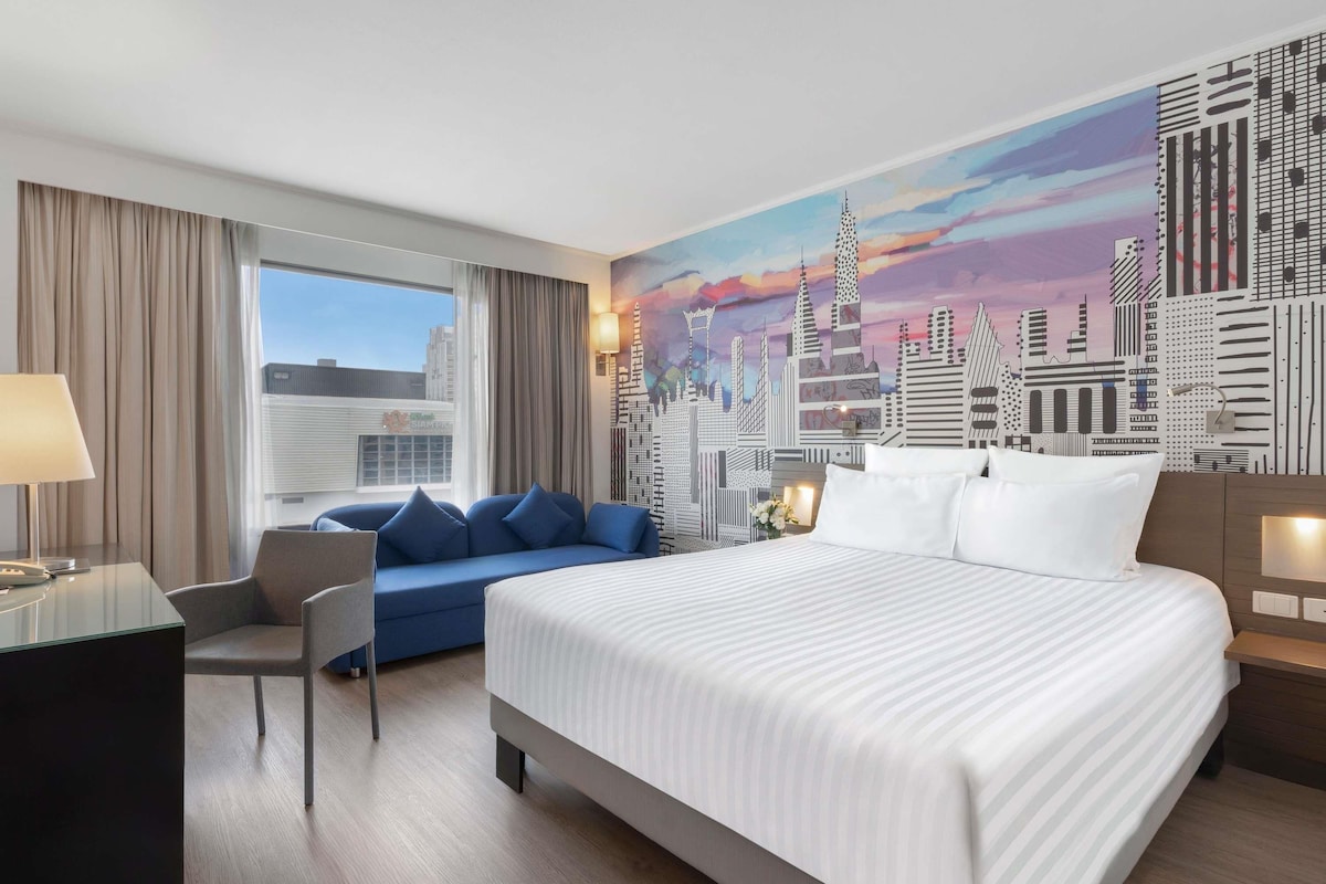 Ein Hotelzimmer mit einem großen Bett, einer Couch, einem Schreibtisch und einem Stadtbild-Wandgemälde.