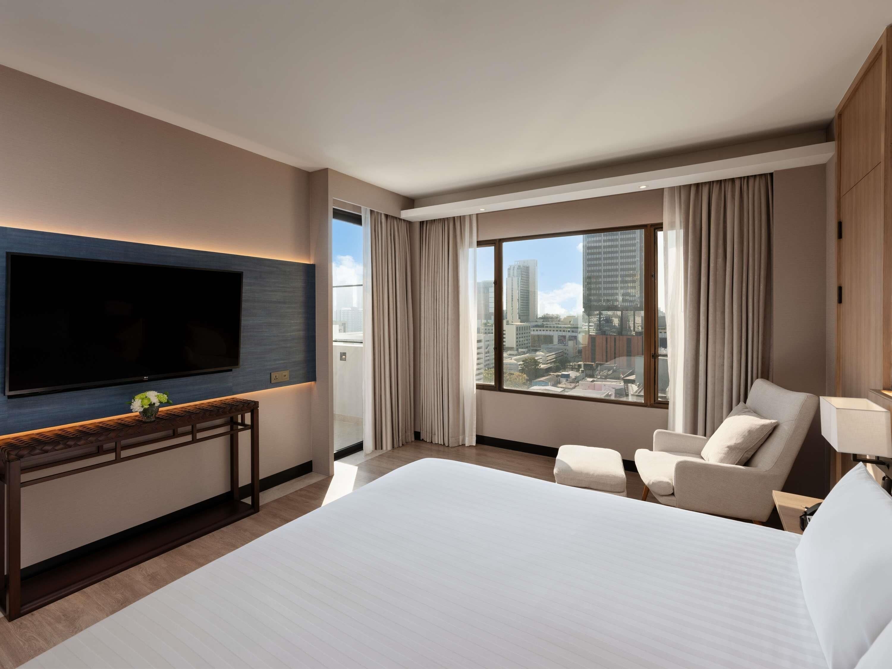 A hotel room with a large bed, a TV mounted on the wall, a desk with a chair, and a view of the city through large windows.