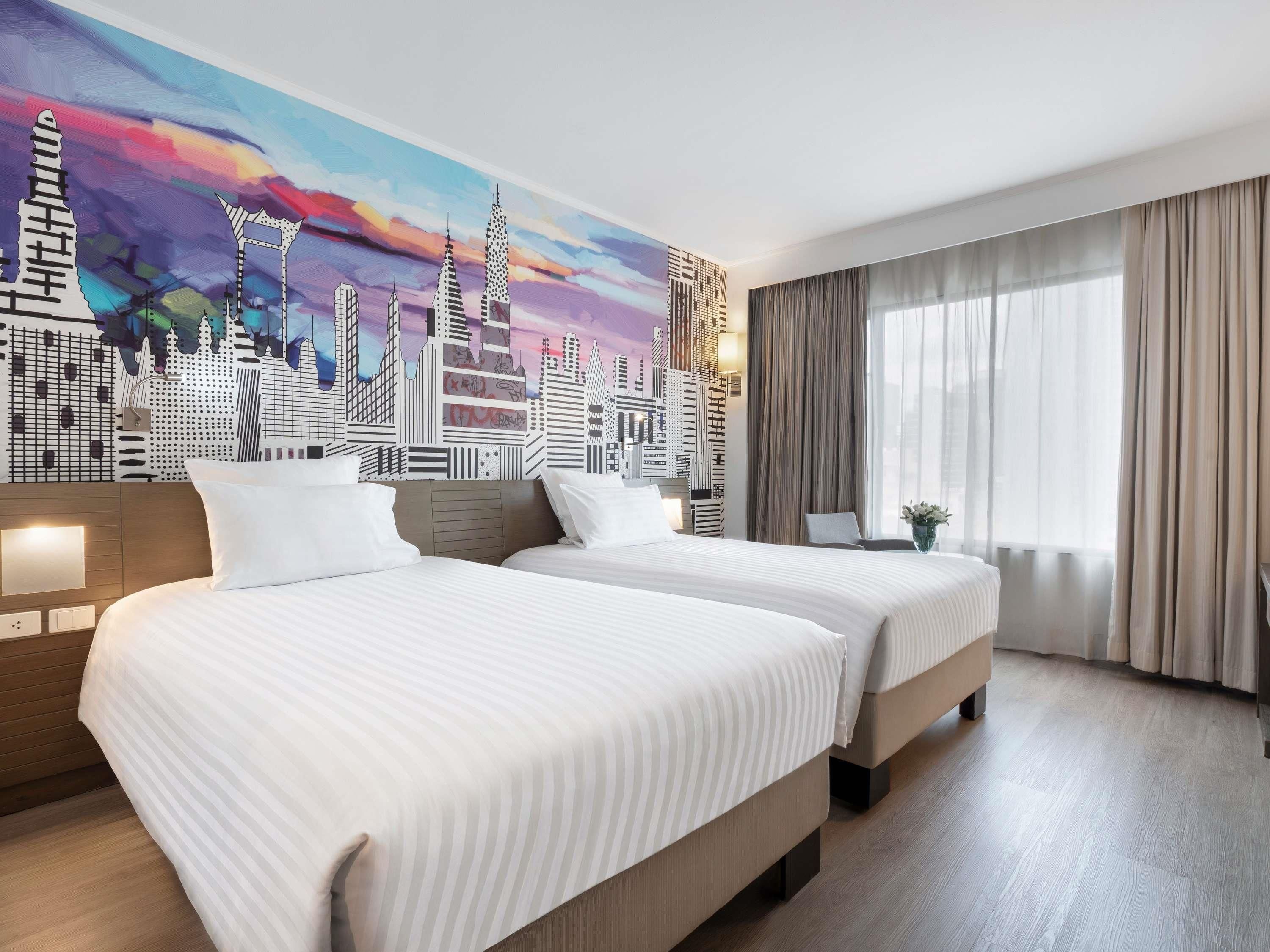 A hotel room with two beds, a cityscape mural, a large window with curtains, and a wooden floor.