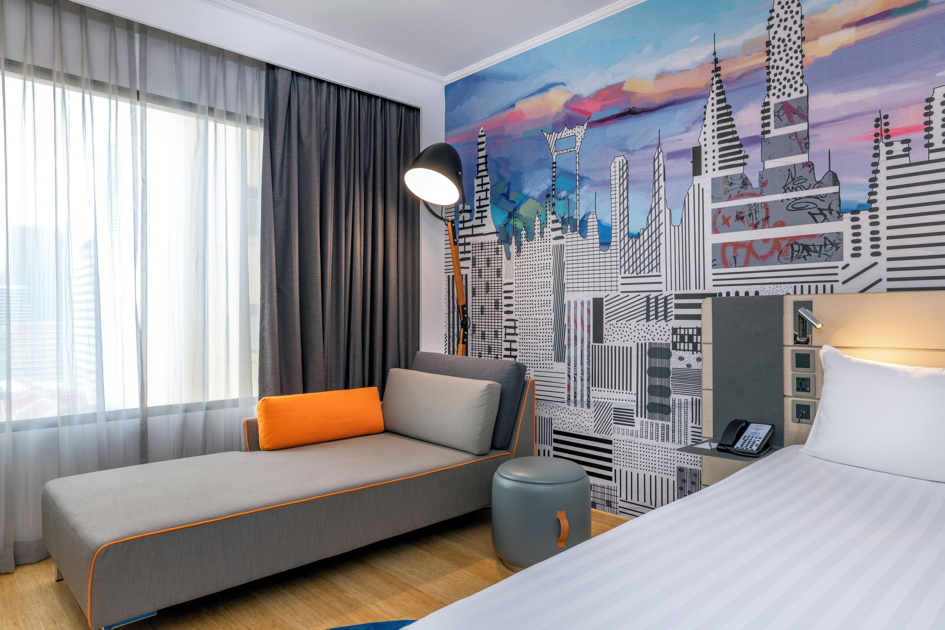 Une chambre d’hôtel moderne avec un canapé gris, un coussin orange, une table de chevet avec une lampe et une fresque murale représentant un paysage urbain.