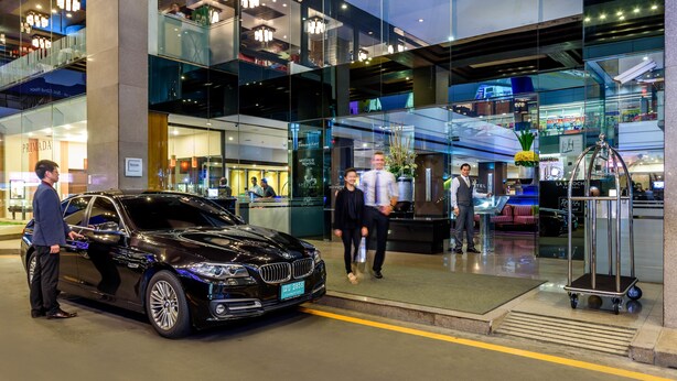 Un moderno vestíbulo de hotel con un coche negro estacionado afuera, paredes de vidrio y una mesa de recepción.