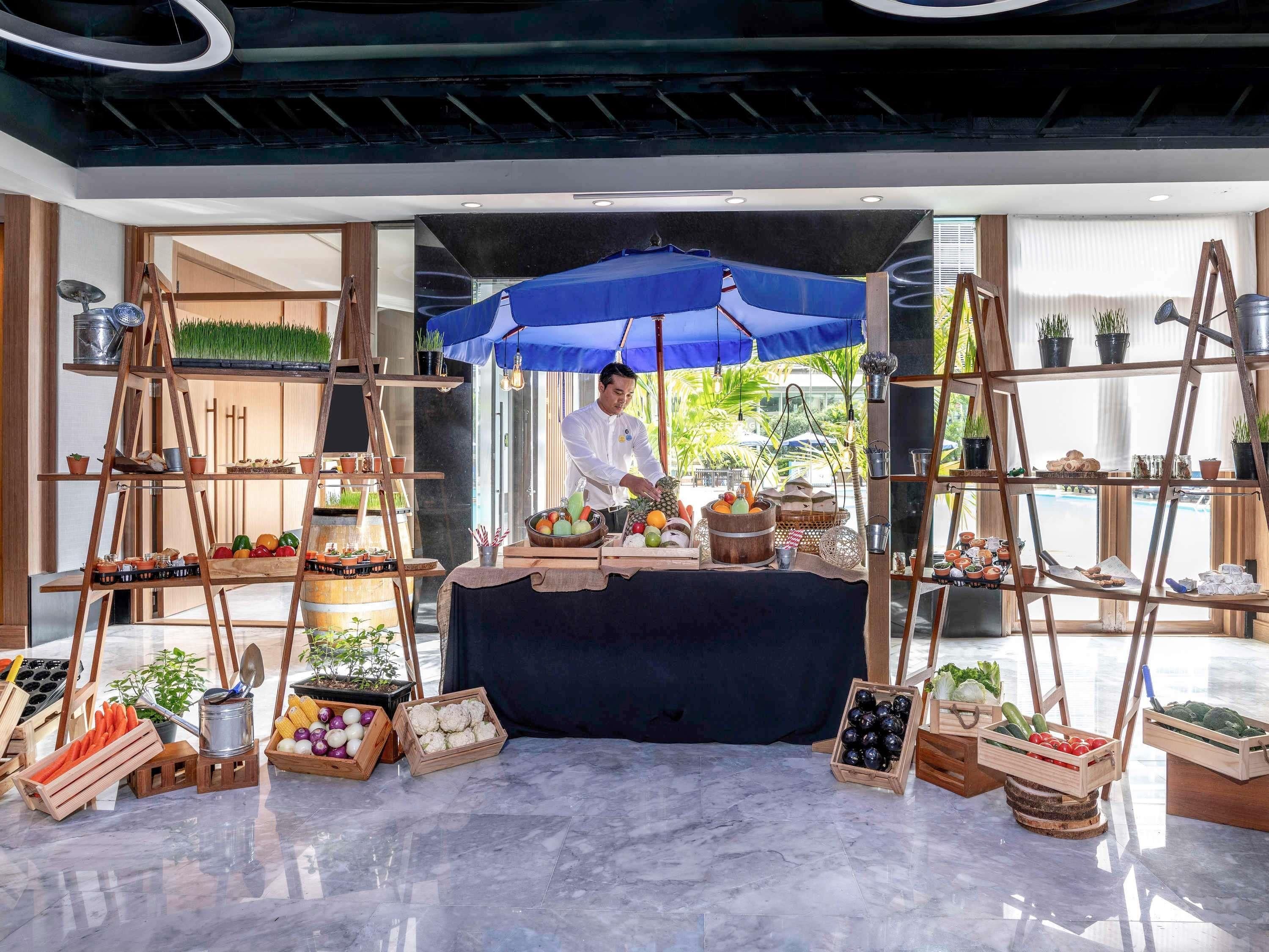 A chef preparing food at a counter with a blue umbrella, surrounded by wooden crates of vegetables and fruits.