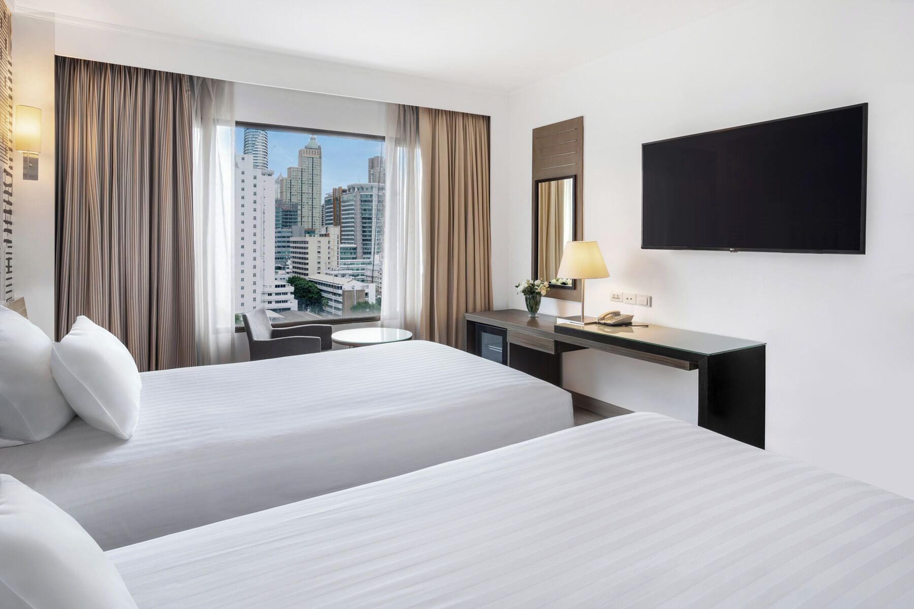 A hotel room with two beds, a desk with a lamp, a flat-screen TV, and a view of city buildings.
