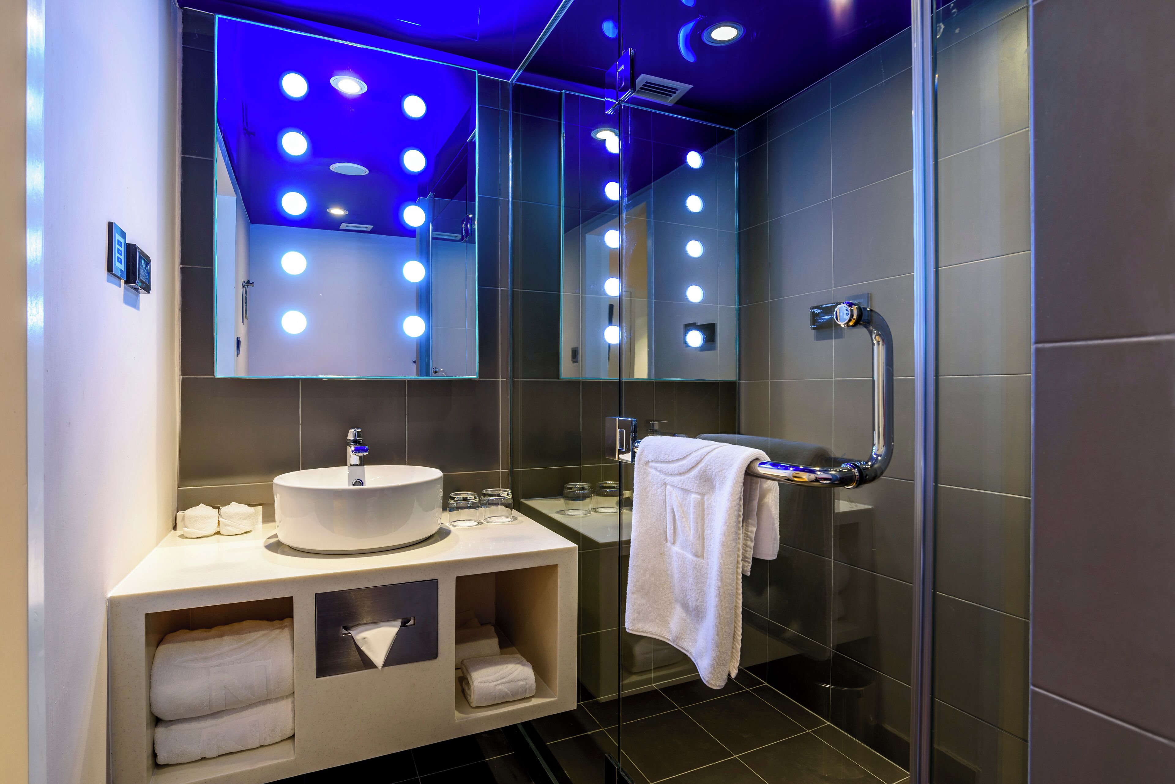 Un baño moderno con un lavamanos redondo, un espejo grande con luces y un box de ducha de vidrio.