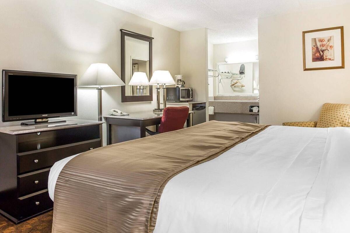 A hotel room with a bed, a desk with a TV, two lamps, a chair, a mirror, and a picture on the wall.