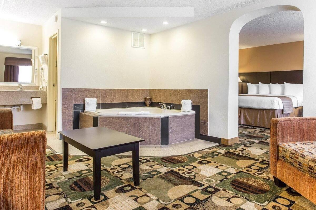 A hotel room with a large bathtub, a separate shower area, a bed with white linens, a small table, and a patterned carpet.