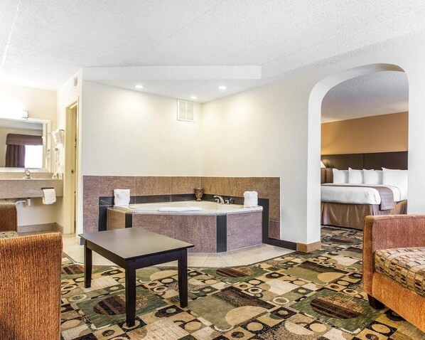 A hotel room with a large bathtub, a separate shower area, a bed with white linens, a small table, and a patterned carpet.