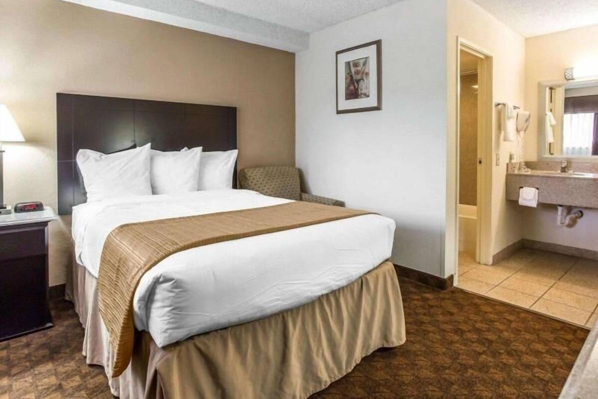 A hotel room with a bed, bedside table, lamp, and a bathroom with a sink and mirror.