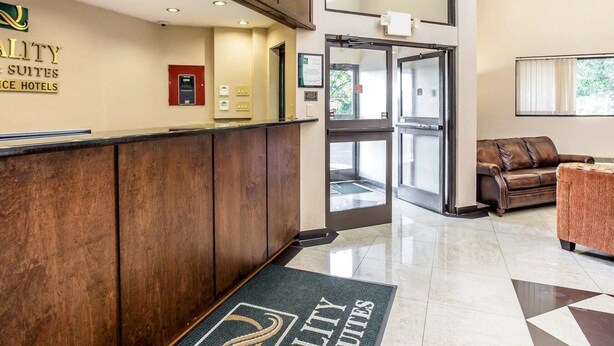 Un espace d’accueil d’hôtel comprenant un comptoir de réception en bois, un kiosque numérique et une porte en verre donnant sur le hall d’entrée.
