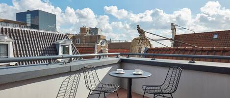 A rooftop terrace with a round table and two chairs, a statue, and a view of surrounding buildings.