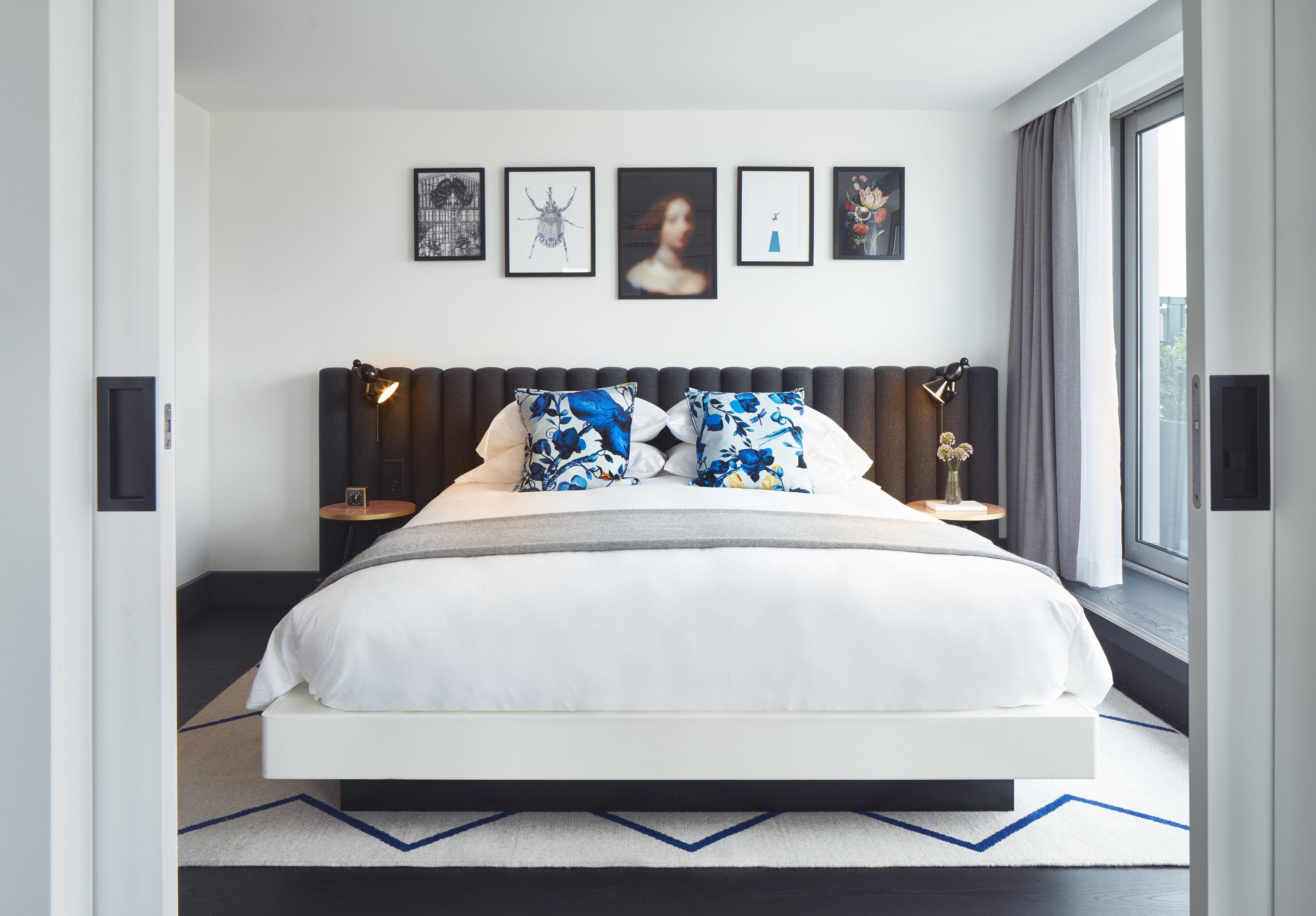A modern bedroom with a large bed, a headboard with integrated lighting, a side table with a lamp, and a window with a view.