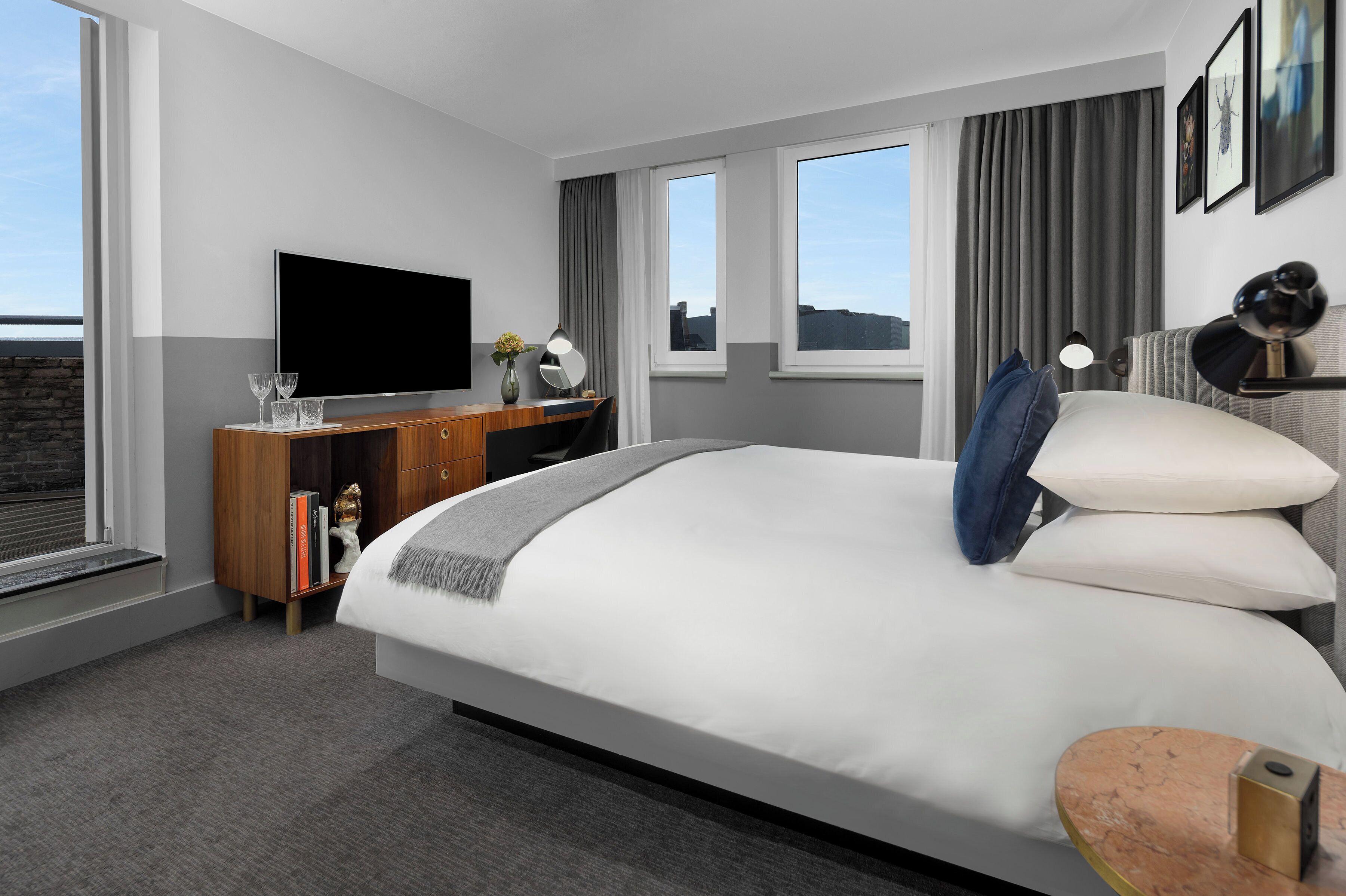 A modern hotel room with a large bed, a flat-screen TV, a wooden desk, and a view of the city through large windows.
