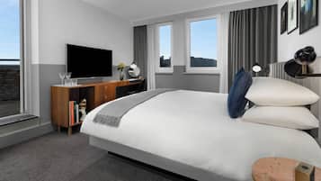 A modern hotel room with a large bed, a flat-screen TV, a wooden desk, and a view of the city through large windows.