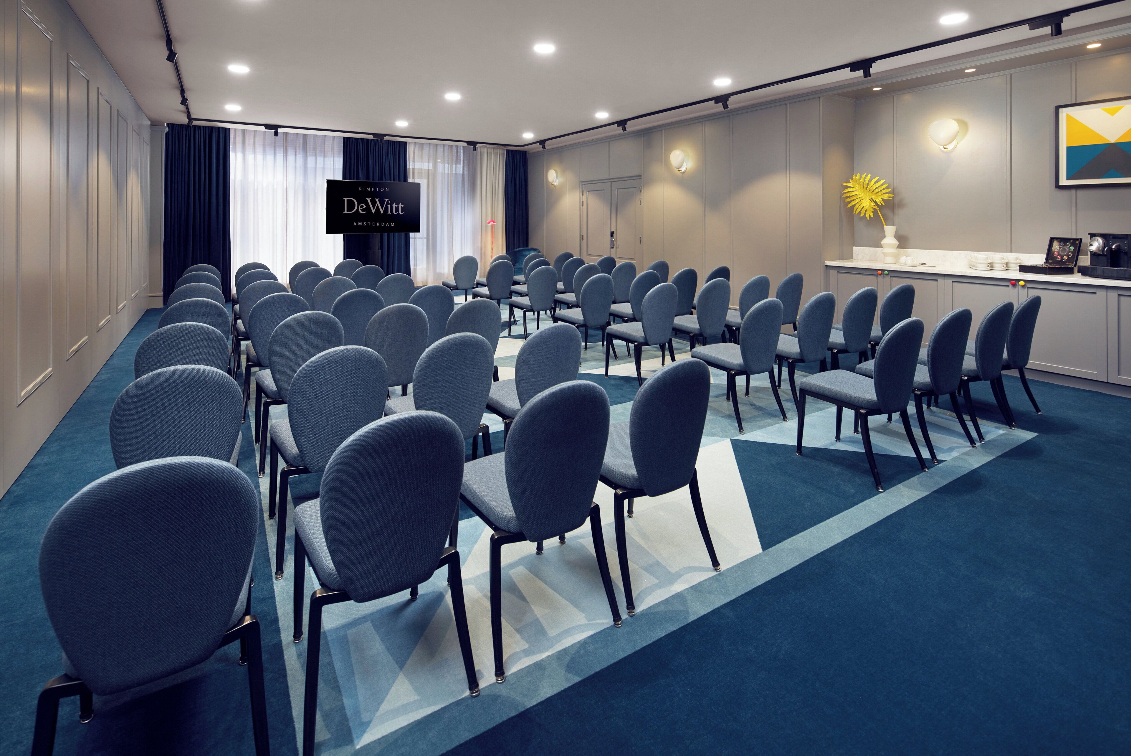 Une salle de conférence avec des rangées de chaises, un pupitre et un espace de réception équipé d’une machine à café.