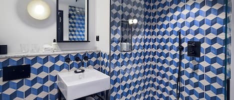 Un baño con pared de diseño geométrico, un lavabo blanco, un espejo y una zona de ducha con mampara de vidrio.