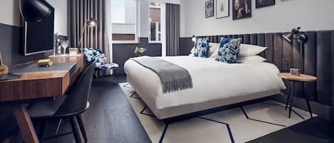 A modern hotel room with a large bed, a desk, and a chair. There are multiple framed pictures on the wall and a window with curtains.