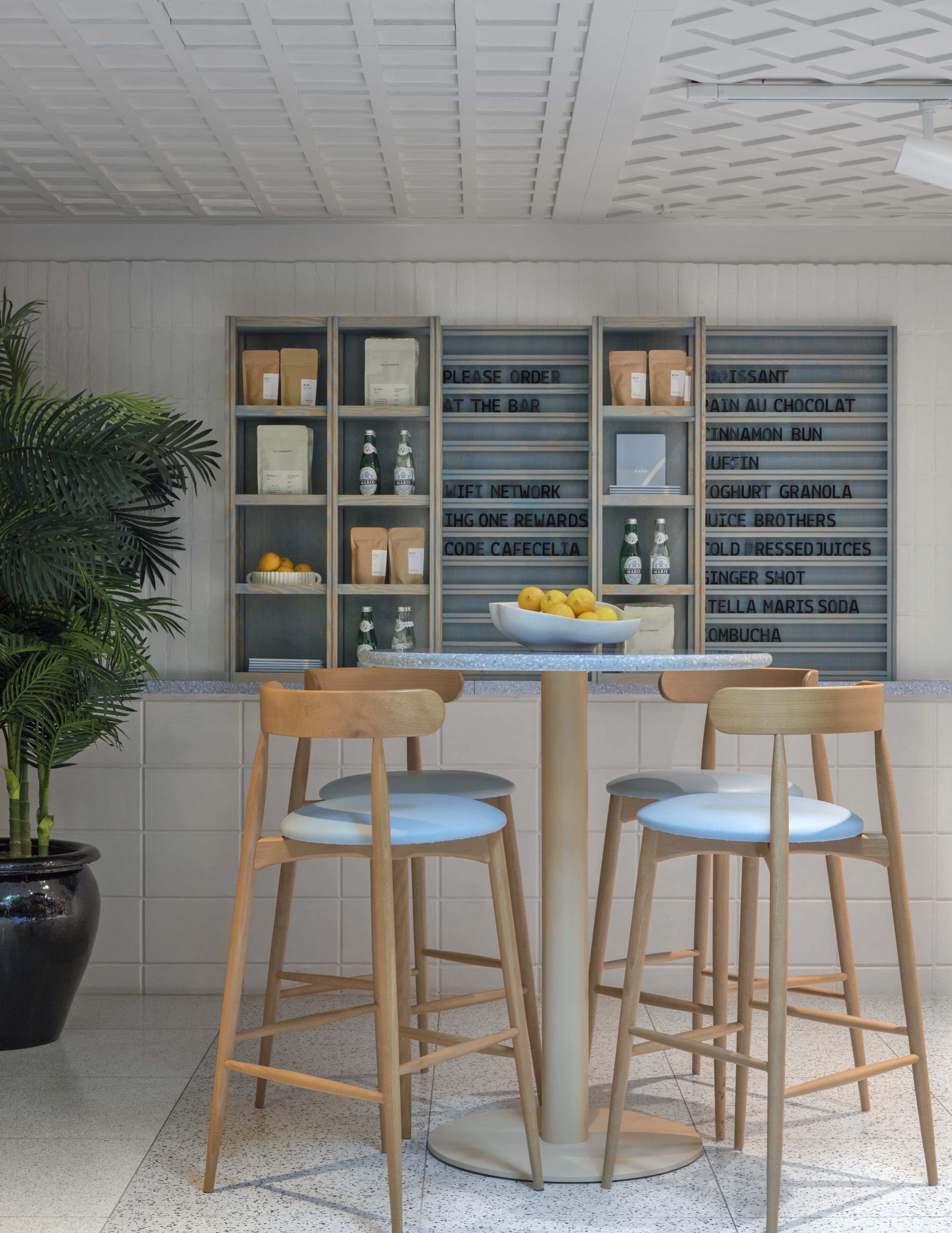 A bar area with wooden stools and a table, a potted plant, and a wall with shelves displaying various items.