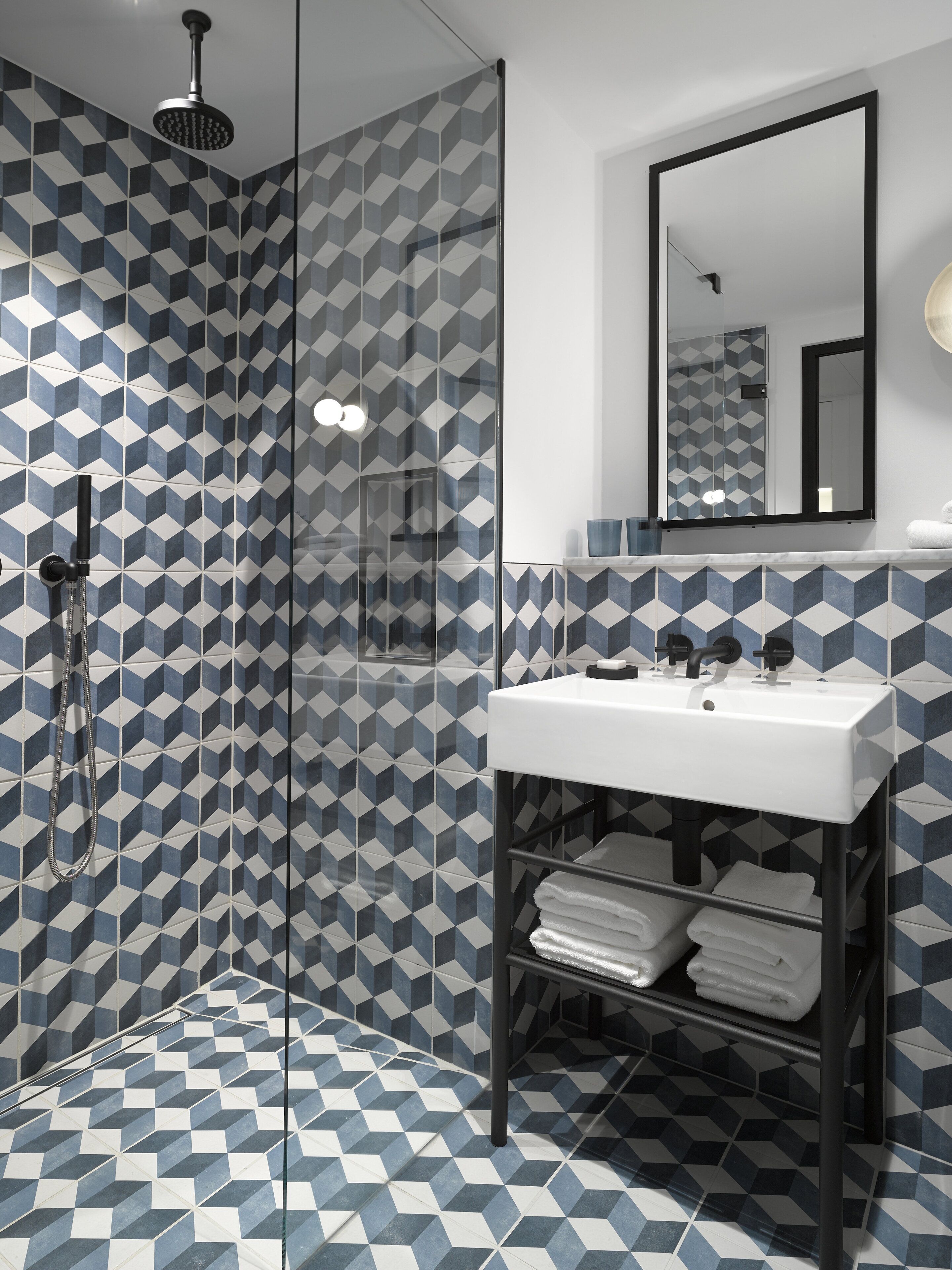 A modern bathroom with a geometric tile pattern, a glass-enclosed shower, a white sink, and a mirror above it.