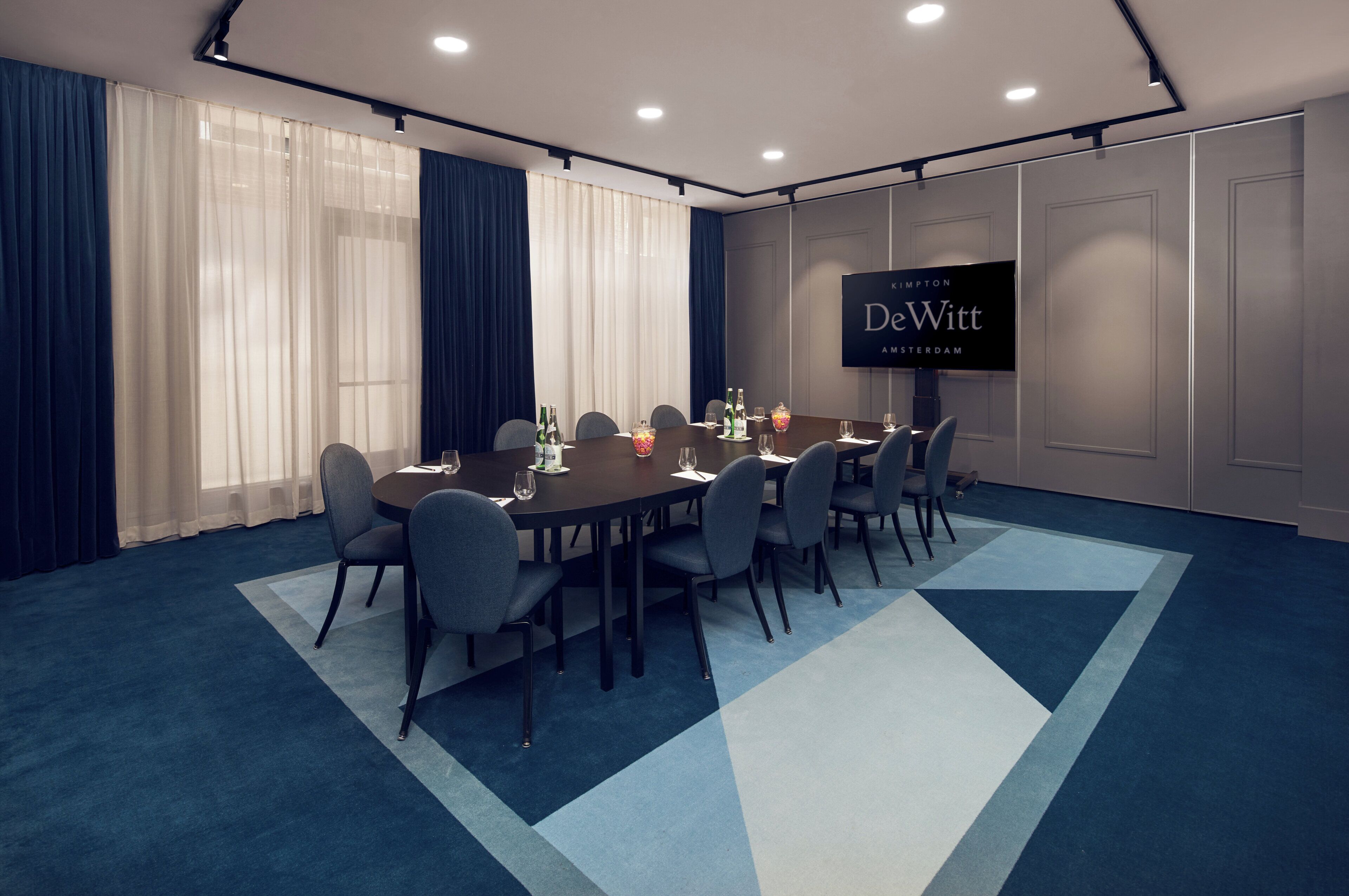 Sala de conferência com uma tela grande exibindo 'DeWitt Amsterdam' e uma mesa montada para uma reunião.