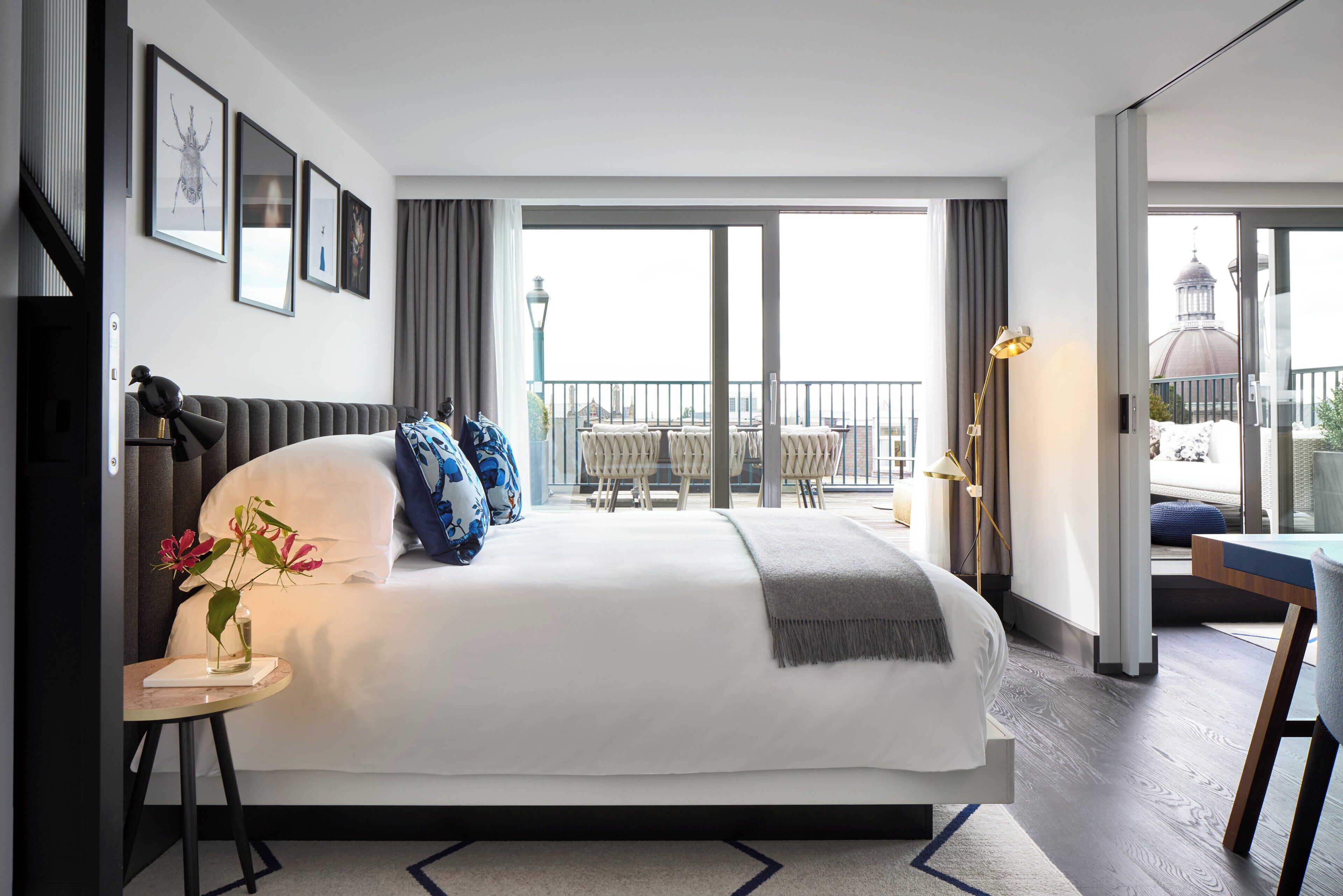 A hotel room with a large bed, a desk with a lamp, a vase with flowers, and a view of the city through the balcony.