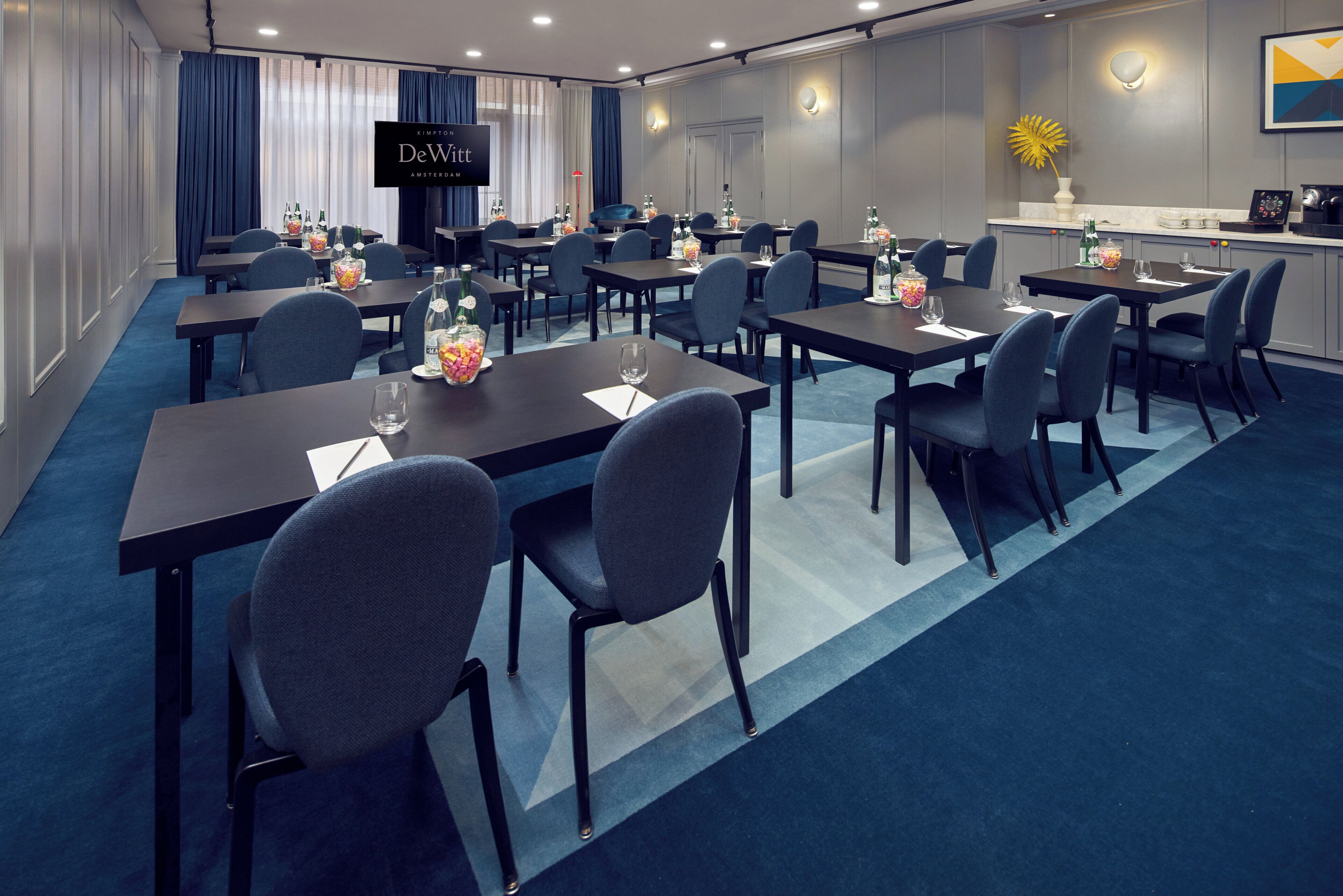 A hotel conference room with tables, chairs, and a television screen displaying 'DeWitt Amsterdam'.