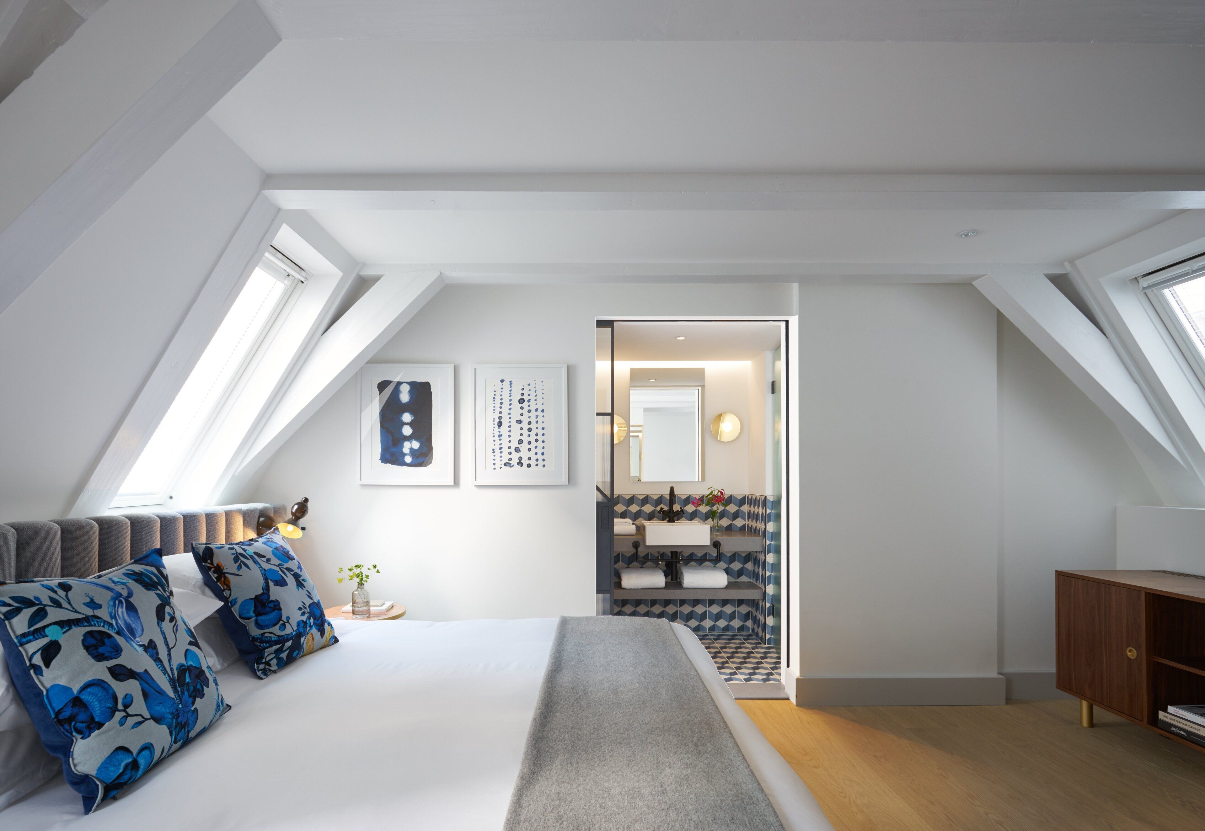 Een moderne slaapkamer met dakraam, een bed met blauwe bloemenkussens, een nachtkastje met vaas, en een houten dressoir.