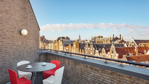 Una terraza en la azotea con pared de ladrillo, una mesa redonda con sillas blancas y rojas, y vista panorámica de la ciudad.
