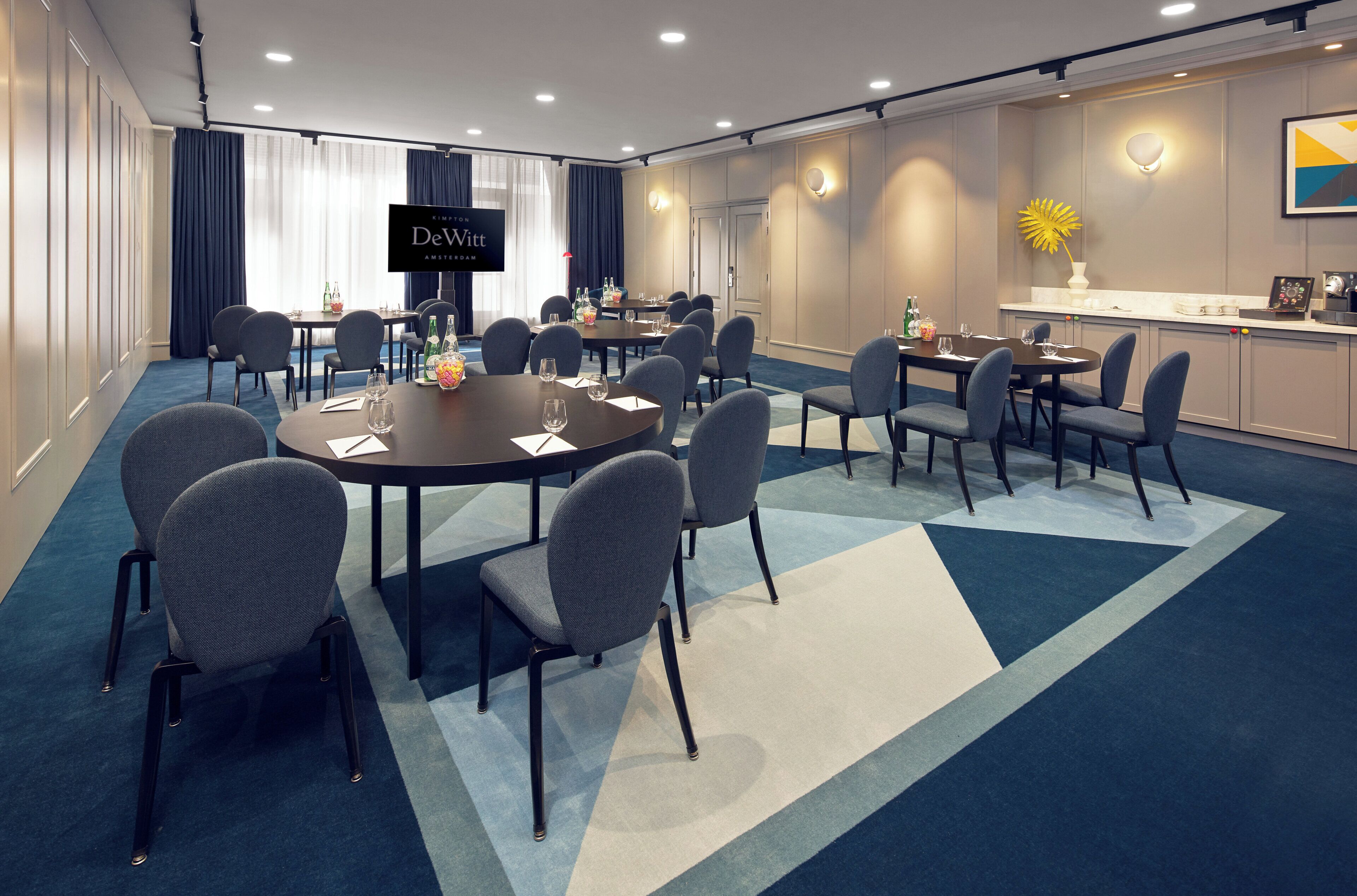 A conference room with a round table, multiple chairs, a television screen, and a kitchenette area.