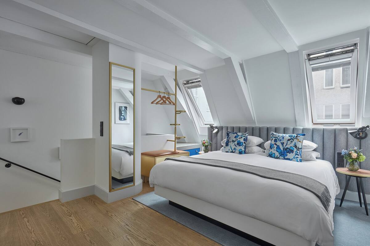 Ein modernes Schlafzimmer mit einem großen Bett, einem Nachttisch, einem Spiegel, einem Kleiderschrank und einem Fenster mit Blick auf Gebäude.