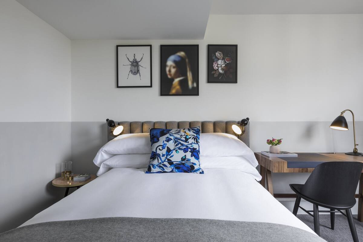 Ein ordentlich eingerichtetes Hotelzimmer mit Bett, Schreibtisch und Stuhl. Darüber an der Wand drei gerahmte Kunstwerke.
