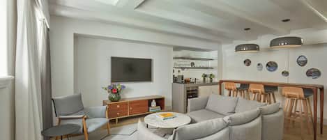 A modern living room with a sofa, a coffee table, and a TV. There is a kitchen area with a dining table and chairs.