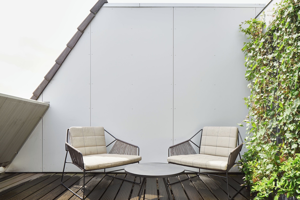 Une terrasse sur le toit, avec deux chaises et une table, entourée d’un mur blanc et d’une plante grimpante.
