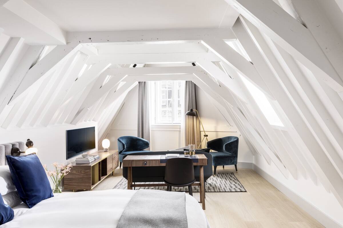 Ein modernes, helles Schlafzimmer im Dachgeschoss mit einem großen Bett, einem Schreibtisch, zwei Stühlen, einem Fernseher und einem Fenster mit Vorhängen.