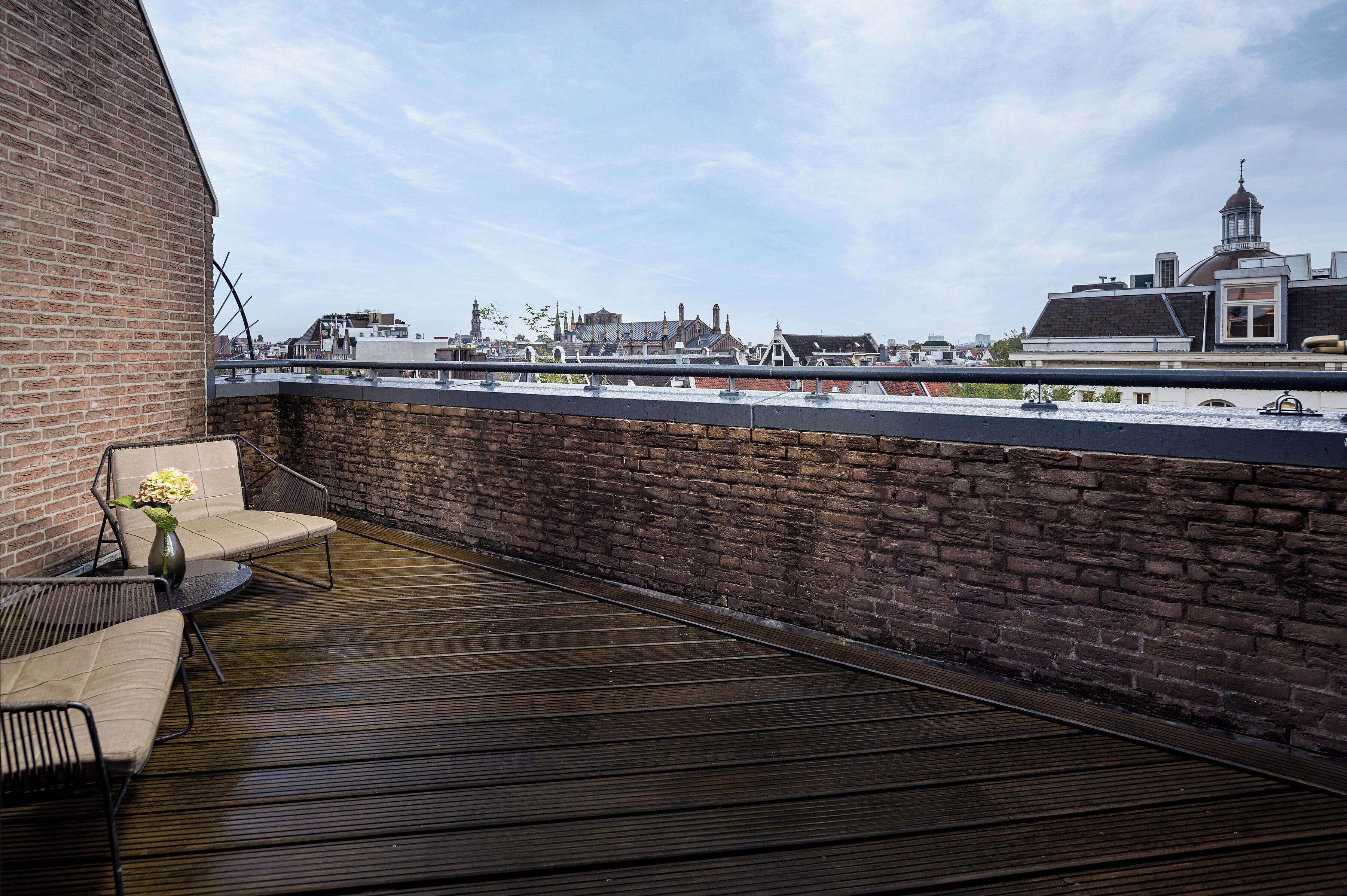 A rooftop terrace with a brick wall, a wooden deck, and a view of buildings in the background.