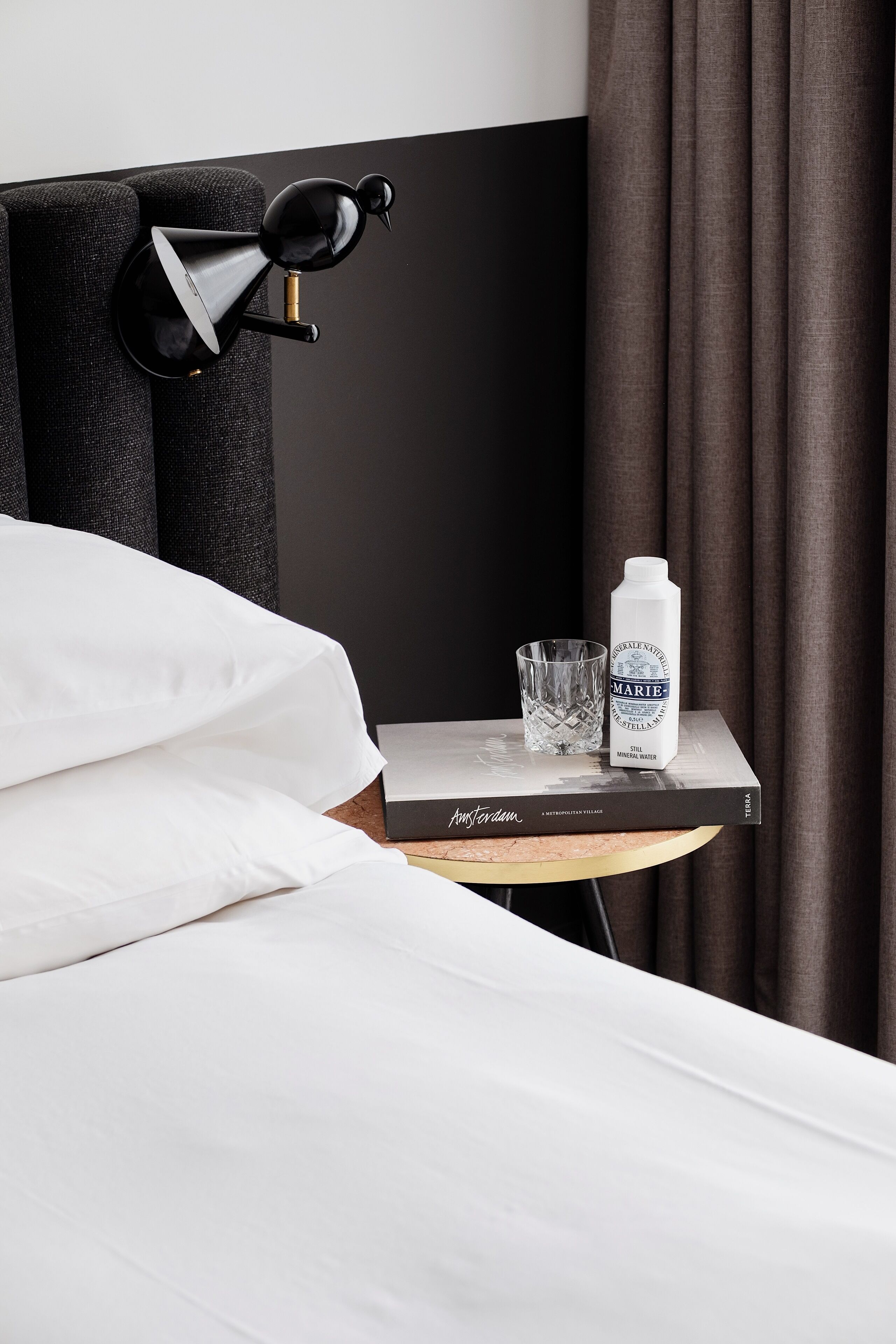 Una cama bien hecha con sábanas blancas, una lámpara negra sobre una mesita de noche, una botella de agua y un vaso.