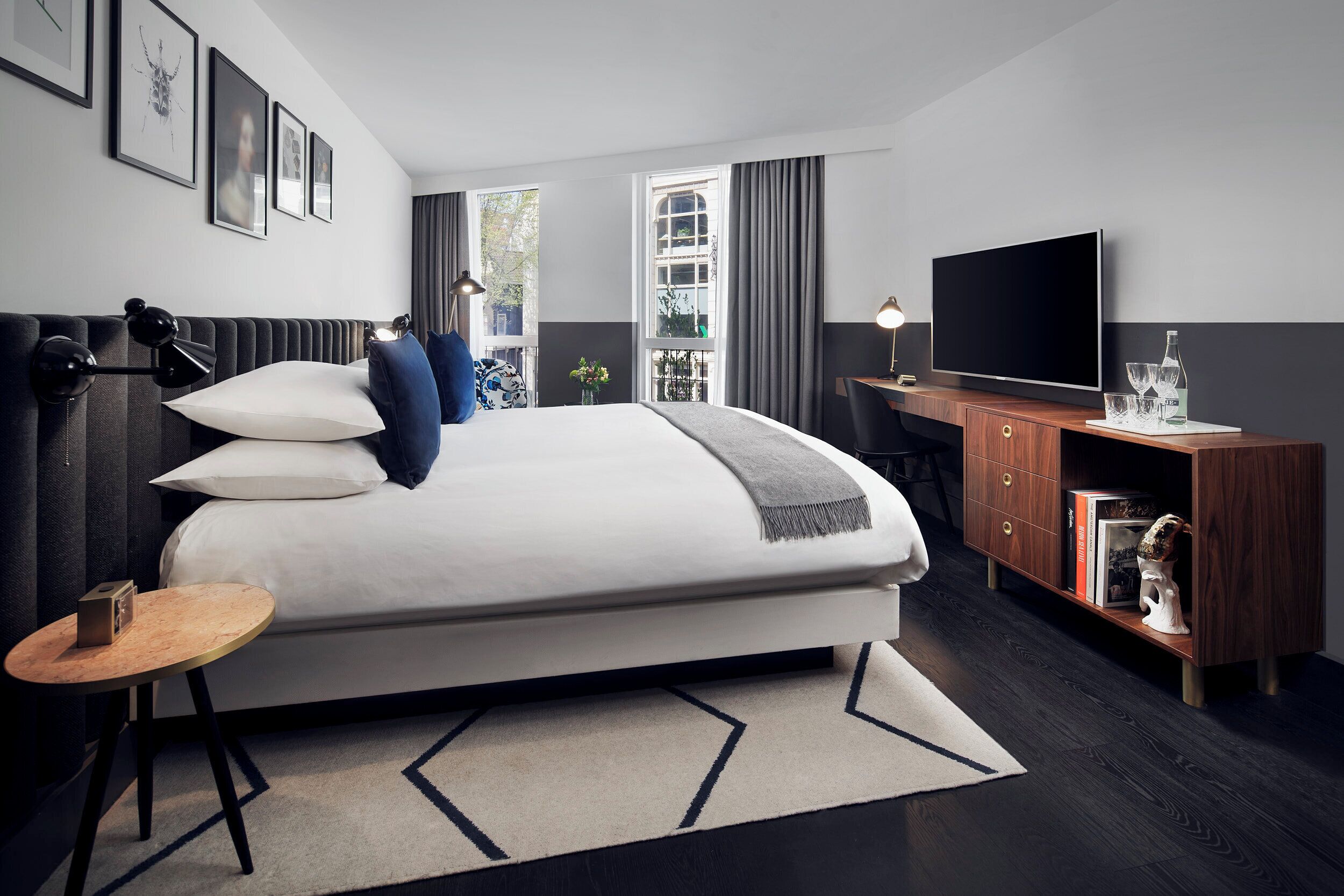 A modern hotel room with a large bed, a desk with a flat-screen TV, a small side table, and a window with curtains.