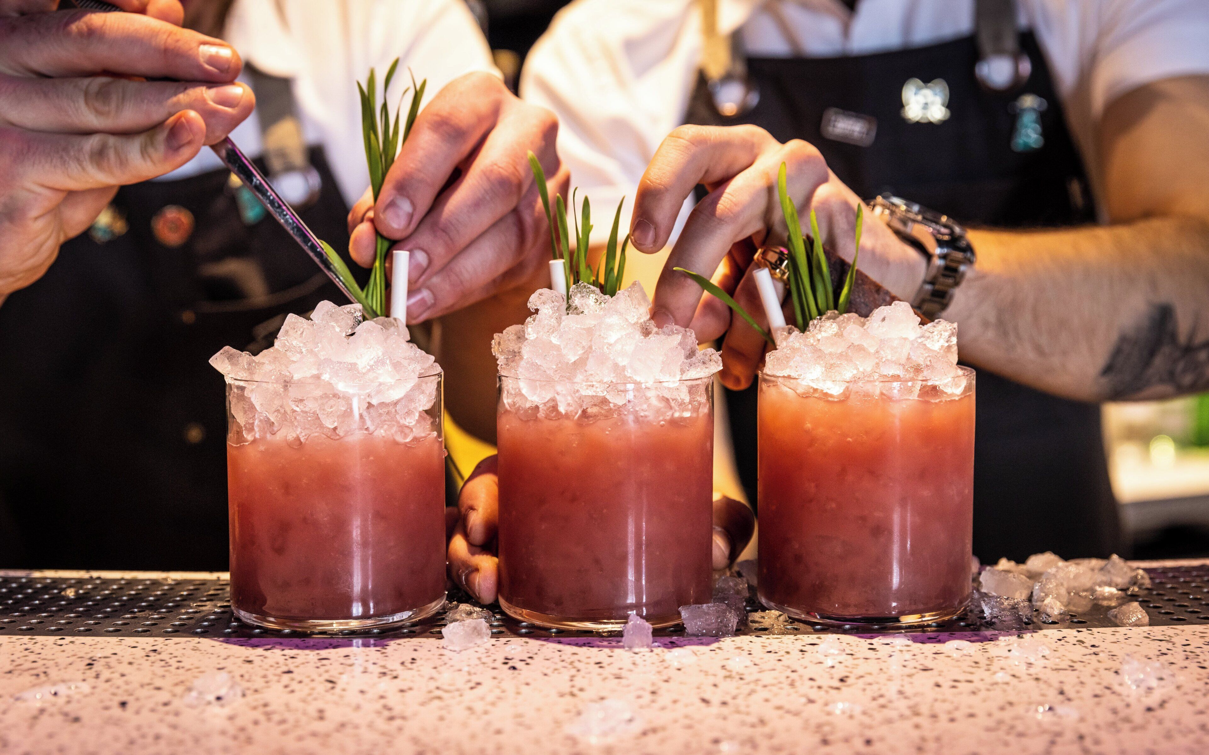 Three glasses of a red beverage with ice and garnished with green herbs, placed on a speckled countertop.