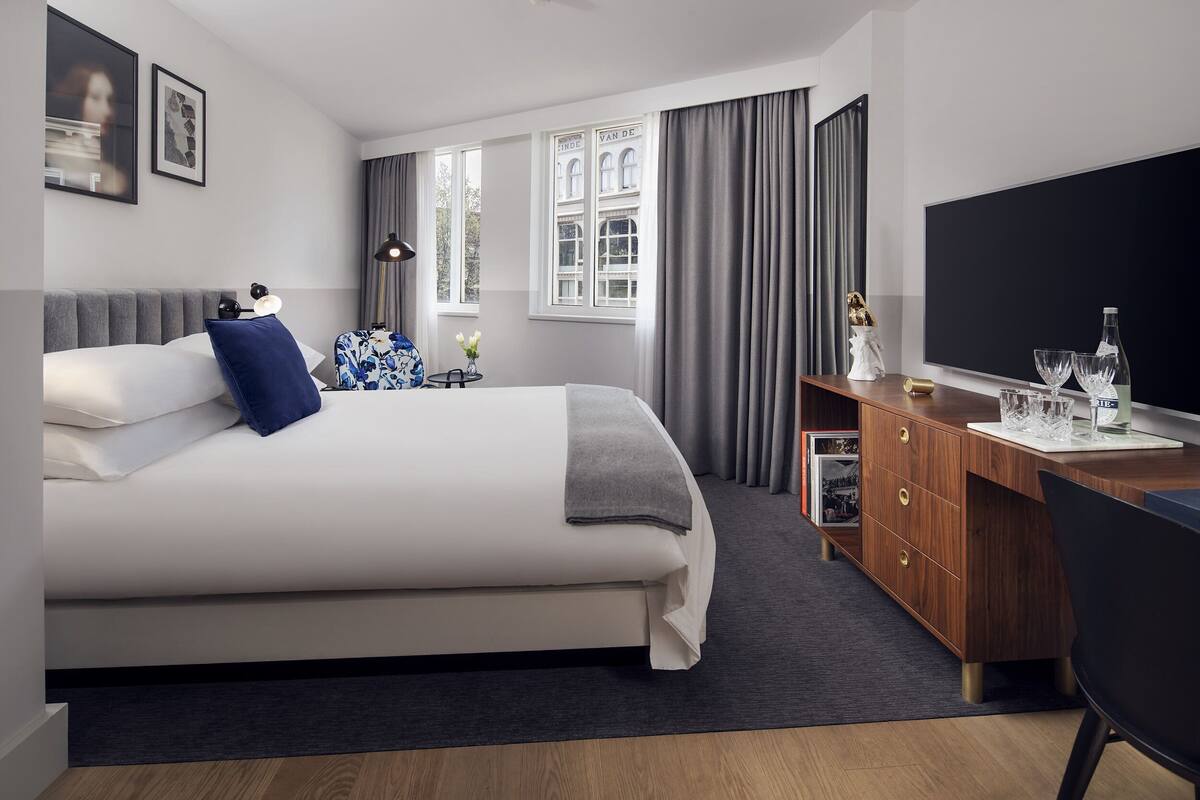 Ein modernes Hotelzimmer mit einem großen Bett, einem Holztisch, einem Flachbildfernseher und Blick auf ein Gebäude durch das Fenster.
