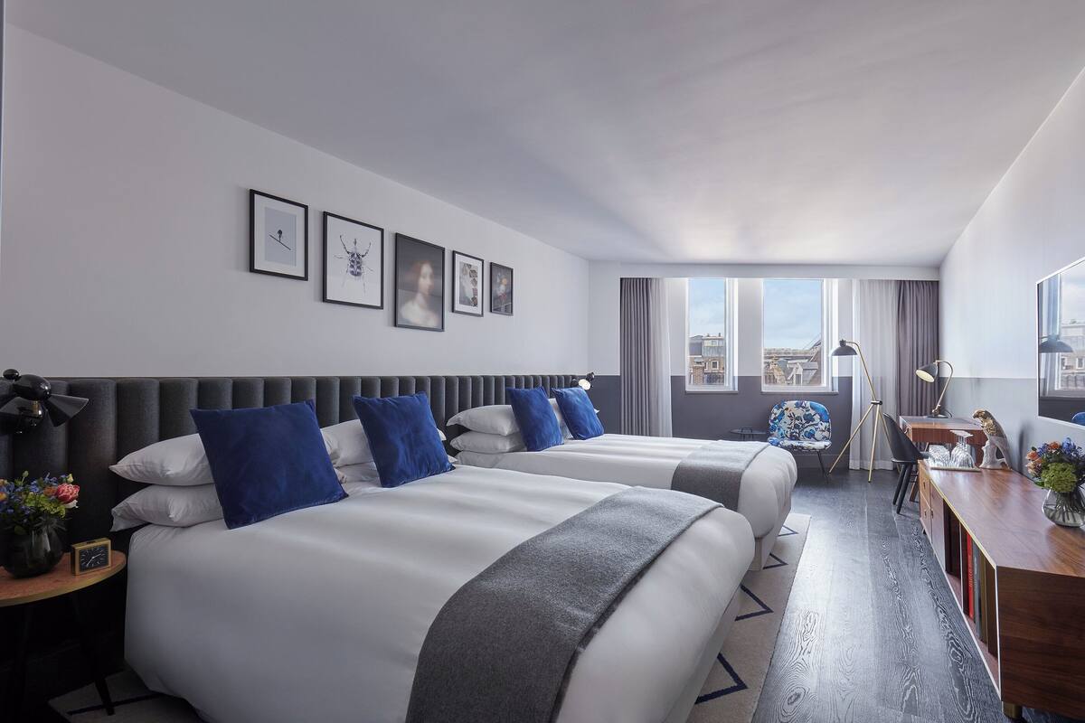 Ein Hotelzimmer mit zwei Betten, einem Schreibtisch, einem Stuhl und einem großen Fenster mit Blick auf die Stadt.
