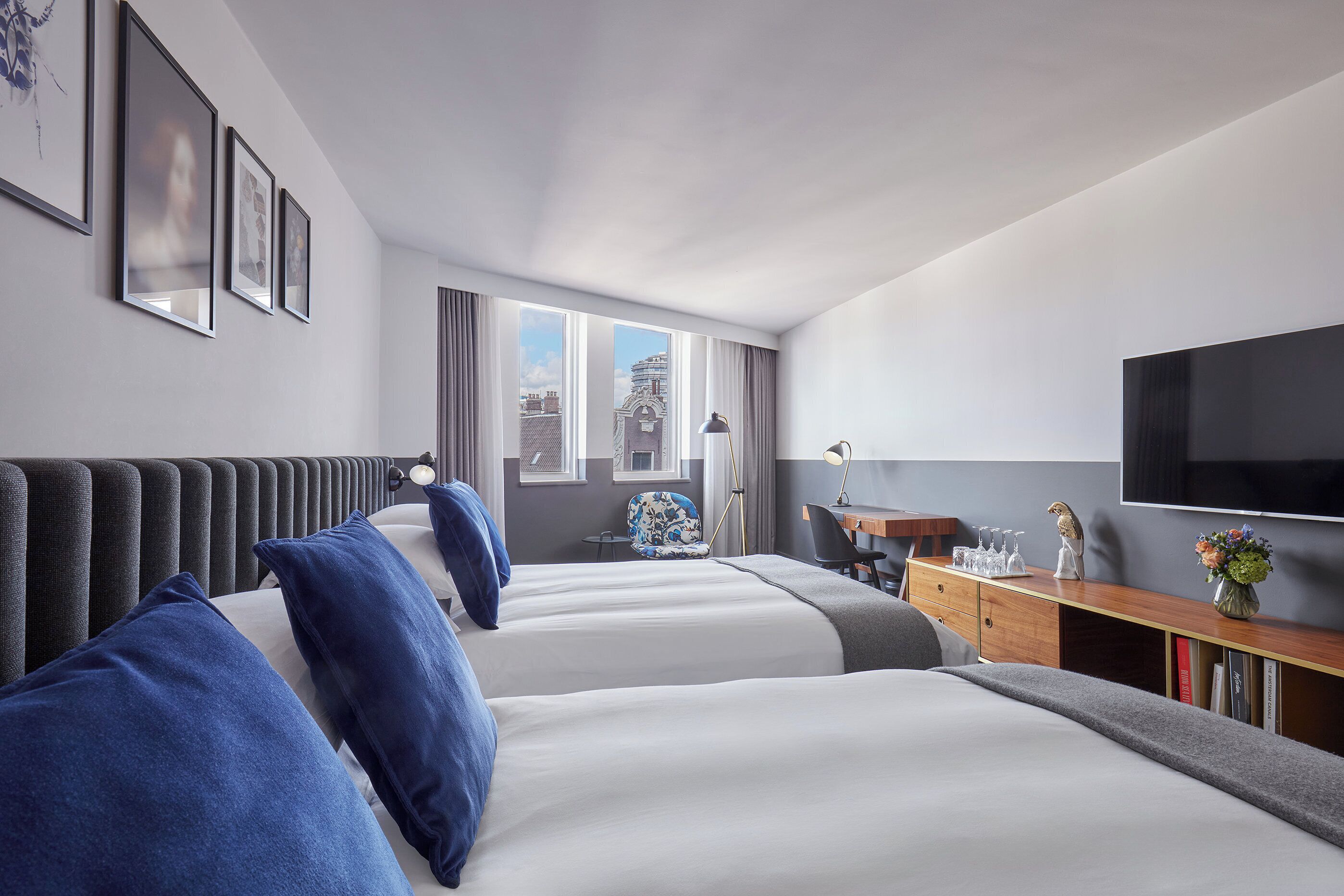 A hotel room with two beds, a desk, a TV, and a window with a city view.