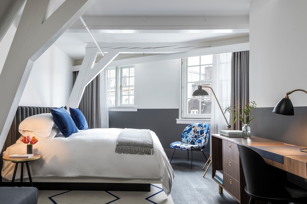 Une chambre moderne avec un lit, une table de chevet, une lampe, une chaise, un bureau et une vue sur la ville par la fenêtre.