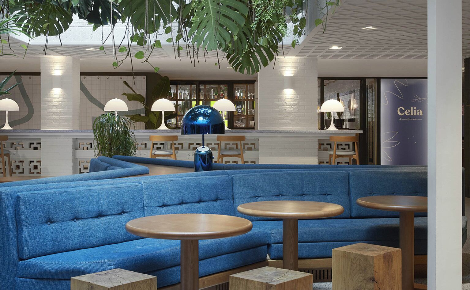 Een moderne lounge met blauwe zitplaatsen, ronde houten tafels en hangplanten.