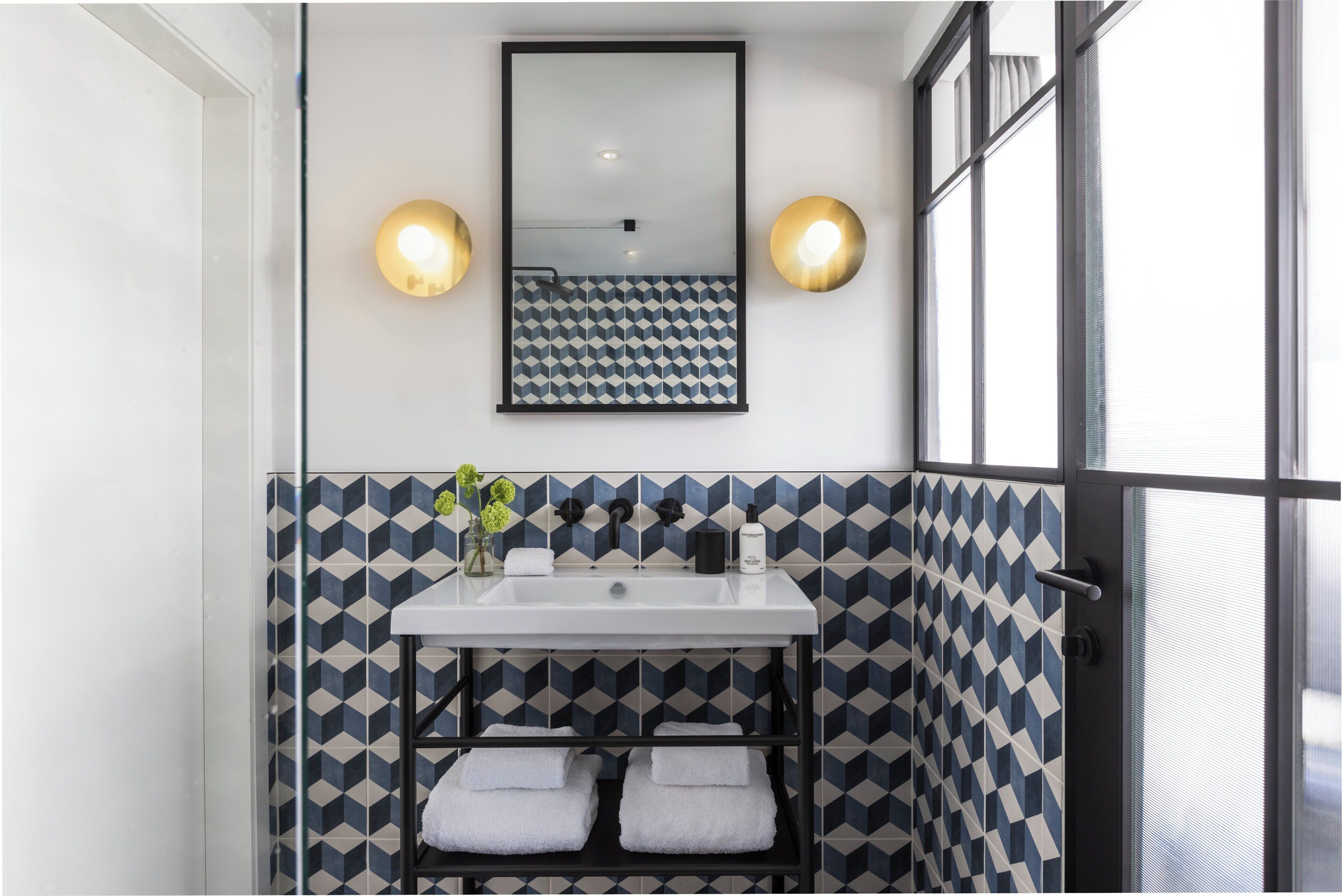 A bathroom with a geometric patterned tile backsplash, a white sink, two wall-mounted lights, and a window with blinds.