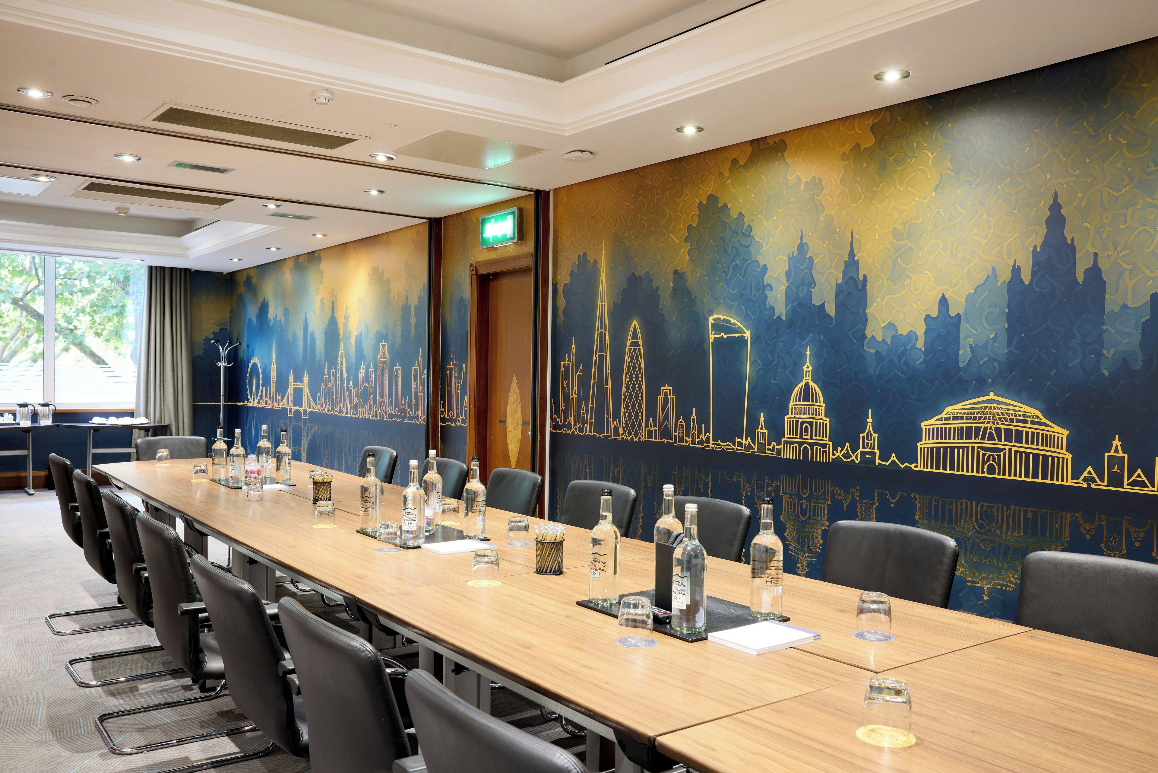 Un salón de conferencias con una larga mesa, sillas y un mural con un paisaje urbano en la pared.