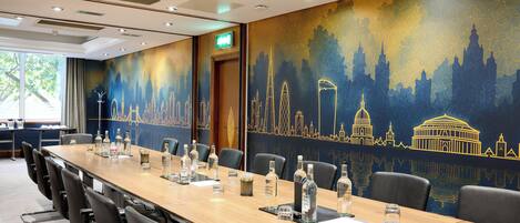 Un salón de conferencias con una larga mesa, sillas y un mural con un paisaje urbano en la pared.