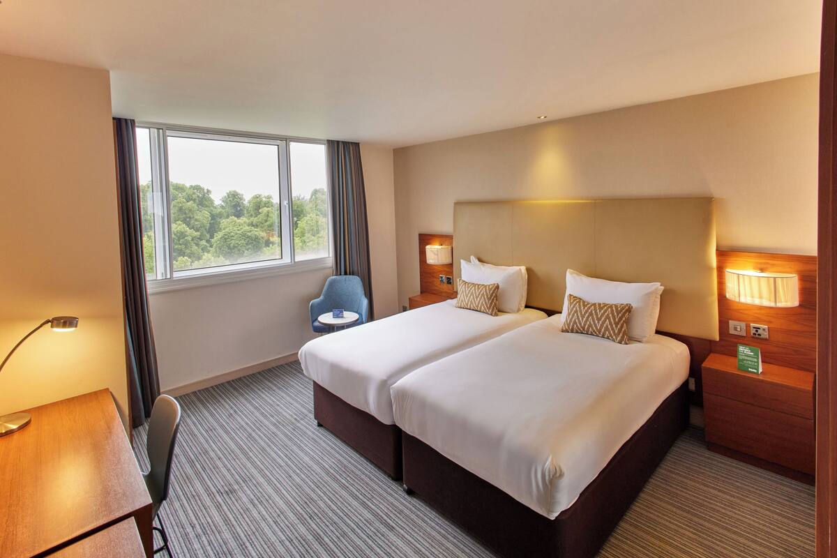 A hotel room with two beds, a desk, a chair, a window with curtains, and a nightstand with a lamp.