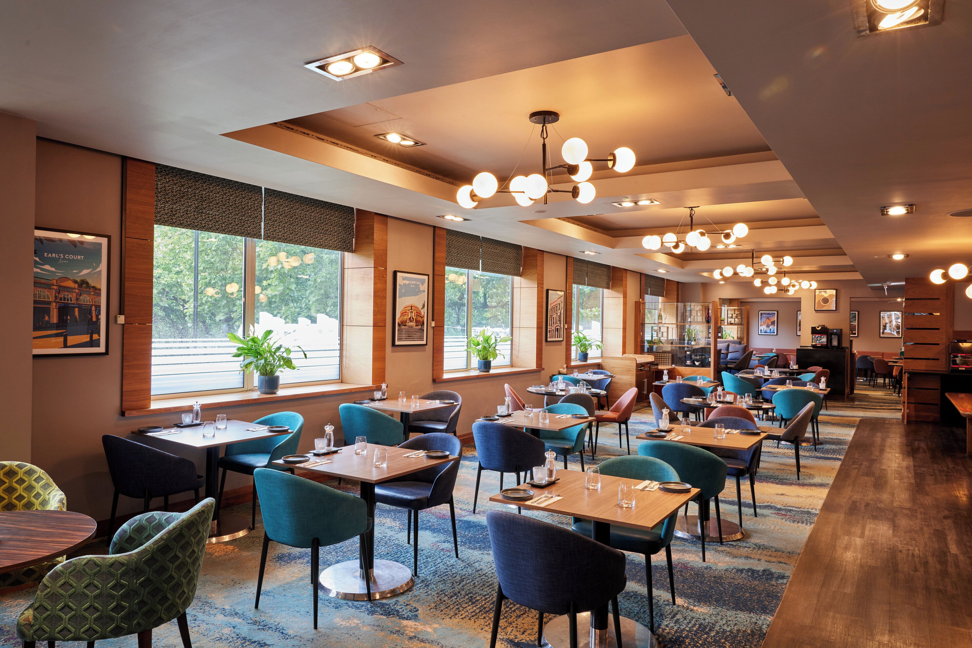 A spacious restaurant with multiple tables and chairs, large windows with plants, and a modern ceiling design.