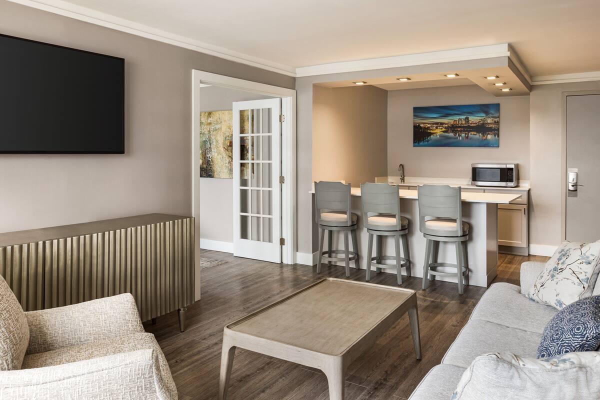 En moderne stue med sofa, sofabord og et tv ophængt på væggen. Der er et køkkenområde med spisebord og barstole.