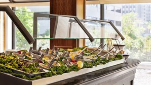 A buffet with various food items and utensils, set against a backdrop of large windows and a view of buildings and trees. - Crowne Plaza Knoxville Downtown University by IHG (Knoxville)