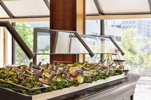 A buffet with various food items and utensils, set against a backdrop of large windows and a view of buildings and trees. - Crowne Plaza Knoxville Downtown University by IHG (Knoxville)